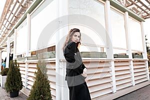 Pretty attractive modern young woman in a chic long black coat in posing near a summer wooden house in the city.
