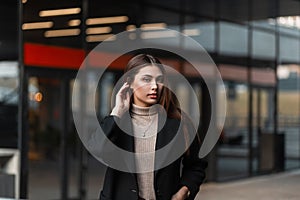 Pretty attractive young woman fashion model straightens hair outdoors in a spring day. Beautiful cute girl in a stylish black coat