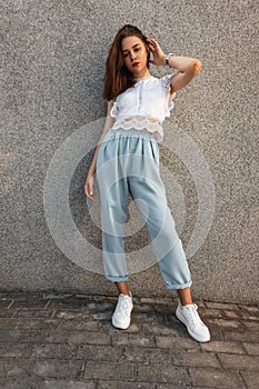 Pretty attractive young woman model in an elegant white lace top in blue vintage pants in fashionable sneakers poses near gray