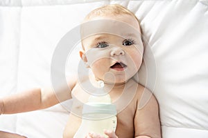 Pretty baby girl drinks milk from bottle lying on bed. Child weared diaper in nursery room.