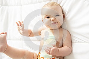 Pretty baby girl drinks milk from bottle lying on bed. Child weared diaper in nursery room.