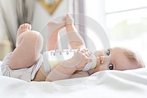 Pretty baby girl drinks water from bottle lying on bed. Child weared diaper in nursery room.