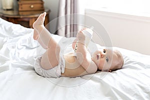 Pretty baby girl drinks water from bottle lying on bed. Child weared diaper in nursery room.