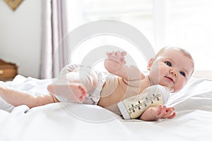 Pretty baby girl drinks water from bottle lying on bed. Child weared diaper in nursery room.