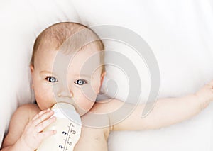 Pretty baby girl drinks water from bottle lying on bed. Child weared diaper in nursery room.