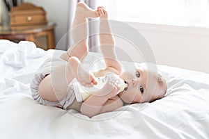 Pretty baby girl drinks water from bottle lying on bed. Child weared diaper in nursery room.