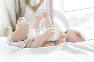 Pretty baby girl drinks water from bottle lying on bed. Child weared diaper in nursery room.