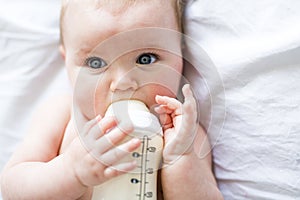 Pretty baby girl drinks water from bottle lying on bed. Child weared diaper in nursery room.