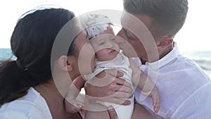 Pretty baby girl at Mediterranean sea beach with father and mother kissing daughter on cheeks. Portrait of charming