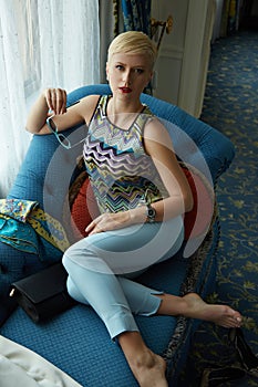 Pretty beautiful woman wear stylish clothes blue pants silk blouse makeup red lipstick blond haircut rest on the sofa hotel room