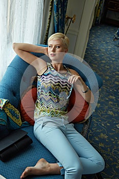 Pretty beautiful woman wear stylish clothes blue pants silk blouse makeup red lipstick blond haircut rest on the sofa hotel room