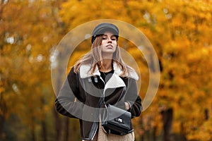 Pretty beautiful young woman in a brown stylish jacket in an elegant hat with a visor with a leather trendy bag posing in the park