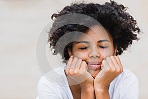 Pretty beautiful young woman with dark skin touching her chin and keeping eyes closed while feeling joyful and excited about
