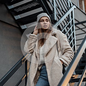 Pretty beautiful young woman model in a fashionable spring vintage knitted hat in an eco fur coat posing standing on an iron gray