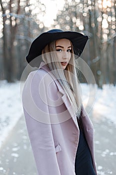 Pretty beautiful young woman in stylish winter glamor clothes goes in a chic hat in a snowy forest on a winter sunny day.