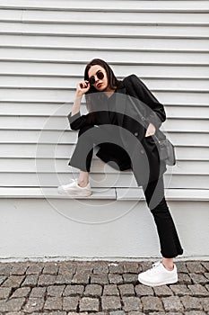 Pretty beautiful young woman in trendy sunglasses in cool black youth clothes in white trendy sneakers with stylish handbag is