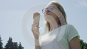 Pretty bespectacled blonde girl eats ice-cream cone thoughtfully looking away from camera in front of blue clear sky