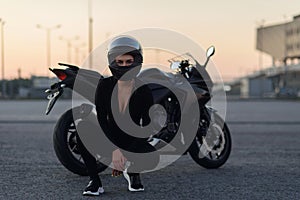 Biker woman in black leather jacket and full face helmet sits near stylish sports motorcycle at urban parking. Traveling