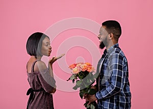 Pretty black lady blowing air kiss to her boyfriend with bouquet of flowers on pink studio background. Valentine's Day