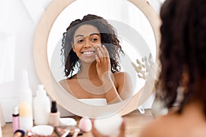 Pretty black woman looking in the mirror and applying facial moisturizer creme photo