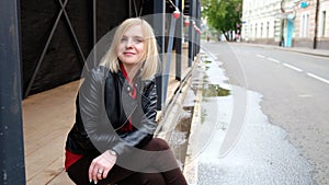 Pretty blond girl in black leather jacket and red shirt sitting outdoor on the street in the city