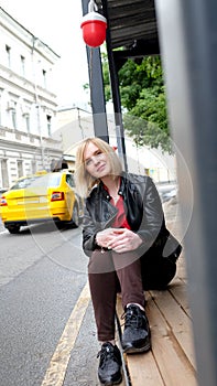 Pretty blond girl in black leather jacket and red shirt sitting outdoor on the street in the city