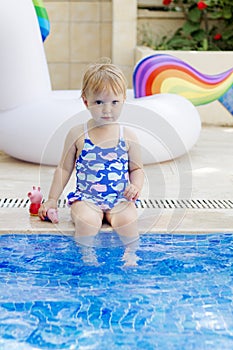 Pretty blond little girl wearing elegant blue swimsuit sitting near the pool in a beautiful sunny summer day