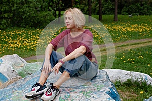 Pretty blond woman sitting on the giant stone in the park, relaxing after long walk, hard day