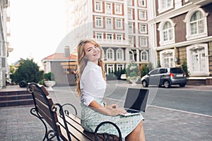 Pretty blonde business woman sitting on banch in city. She holds laptop on knees, smiling to camera.