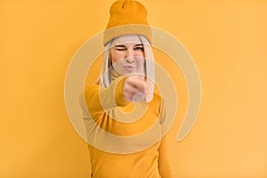 Pretty blonde female gives thumb up, approves new good idea, blink with the eye, wears yellow outfit, stands against yellow studio