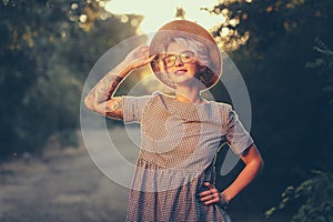 Pretty blonde girl with short haircut is posing and smiling to the camera in forest on sunset background. She wears hat