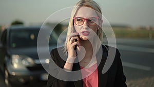 Pretty blonde in glasses talking on a smartphone near a broken car at highway