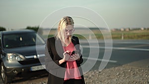 Pretty blonde with glasses typing on a smartphone near a broken car near highway