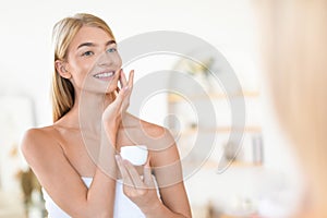 Pretty blonde lady applying moisturizer and enjoying moisturizing skincare indoors