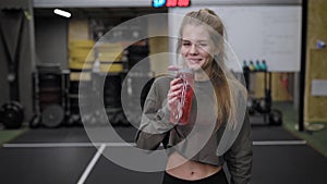 Pretty blonde sporty woman is drinking water from plastic bottle and walking in gymnastic hall in fitness club