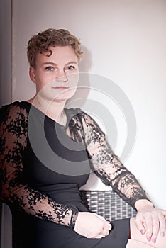 Pretty blonde ukrainian young woman with short hair sitting on a chair, dressed with black dress, against light background