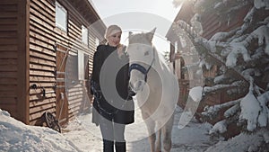 Pretty blonde walks with a beautiful white horse leading her holding a stirrup over a snow-covered country ranch.