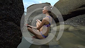 Pretty blonde woman doing yoga between rocks near the sea during beautiful summer day