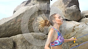 Pretty blonde woman exercising using jumping rope between the rocks near the sea during summer day