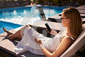 Pretty blonde woman lying in the sun on beach bed with smartphone in her hand near a swimming pool on luxury spa. Girl