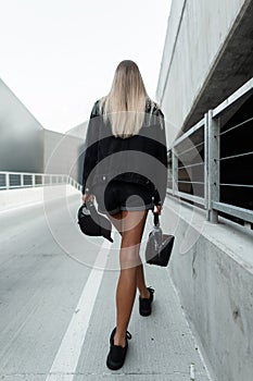 Pretty blonde woman with sexy legs in fashion black jeans outfit with cap and stylish handbag walks in the city, back view