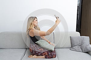 young blonde woman taking photos with her smart phone on the sofa in her living room