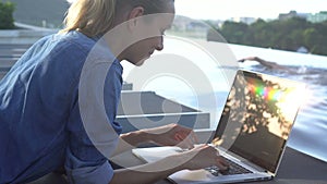 Pretty blonde woman working on a laptop while lying on sunbed with man swimming in infinity pool background