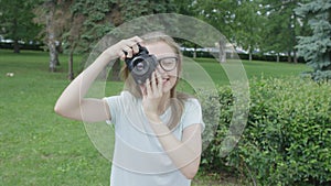 Pretty blonde young woman-photographer in glasses taking pictures in park on a warm sunny day. her camera pointed at
