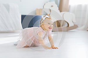 A pretty blue eyed blonde one year old in a pink dress crawls on all fours in the nursery on the floor and smiles fervently