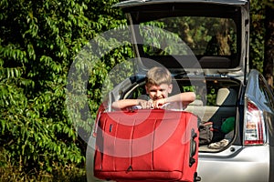 Pretty boy loading the luggage in the trunk of the car. Kid looking forward for a road trip or travel. Family travel by
