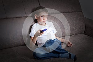 A pretty boy is sitting on the couch using the remote control, the child is watching TV