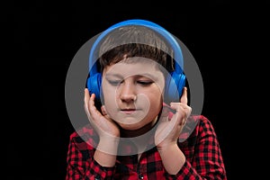 Pretty boy teenager in red plaid shirt attentively enthusiastically listens to music in big blue wireless over-ear headphones