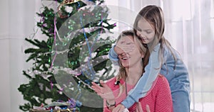 Pretty brunette Caucasian girl closing mother`s eyes with hands and giving her Christmas present. Happy mother receiving