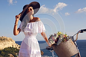 Pretty brunette in elegant dress and hat riding a bicycle along the coast